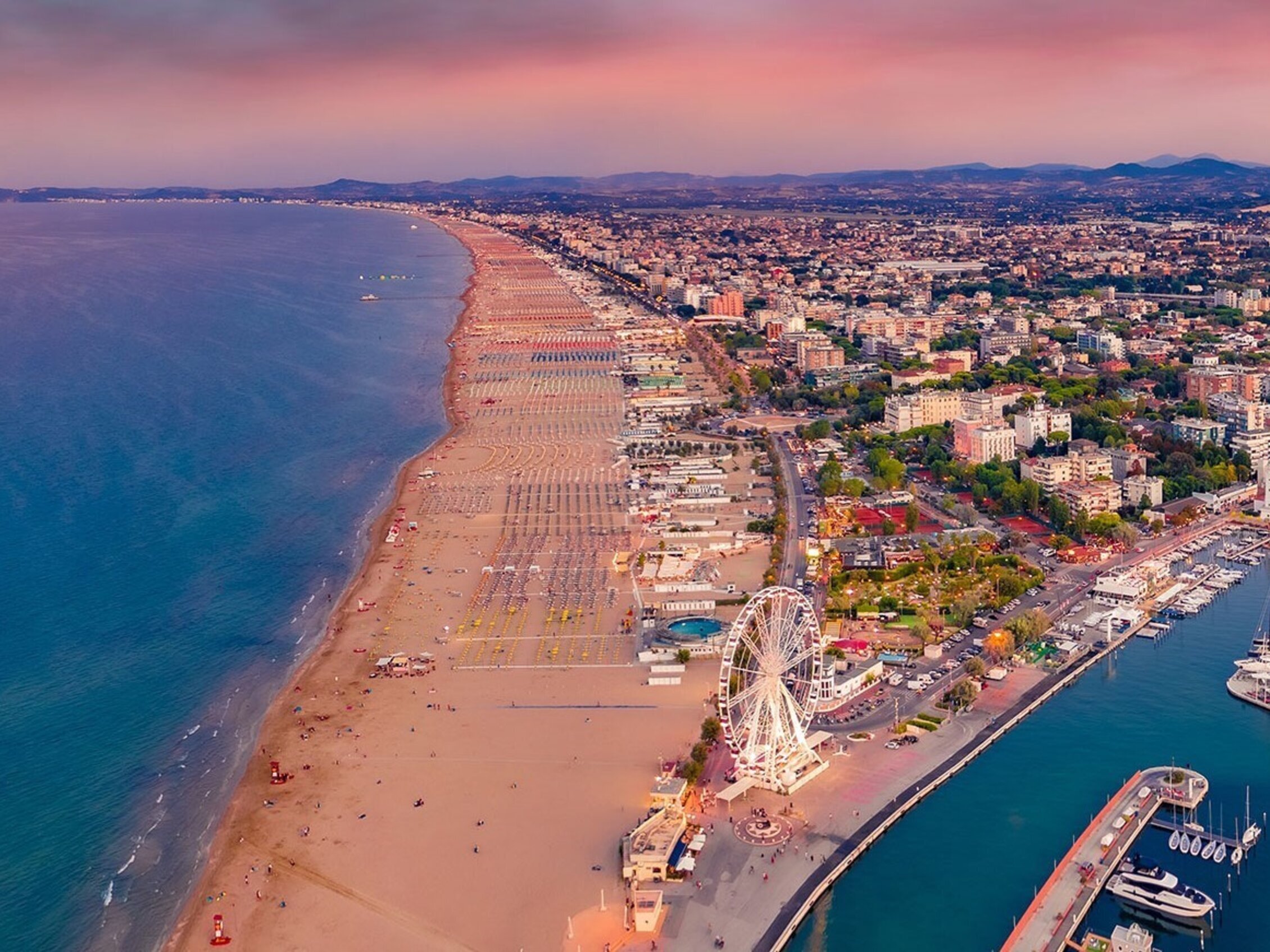 Veduta aerea della spiaggia di Rimini al tramonto, con la lunga distesa di sabbia affacciata sull’Adriatico, la ruota panoramica vicino al porto turistico e la città che si estende nell’entroterra.