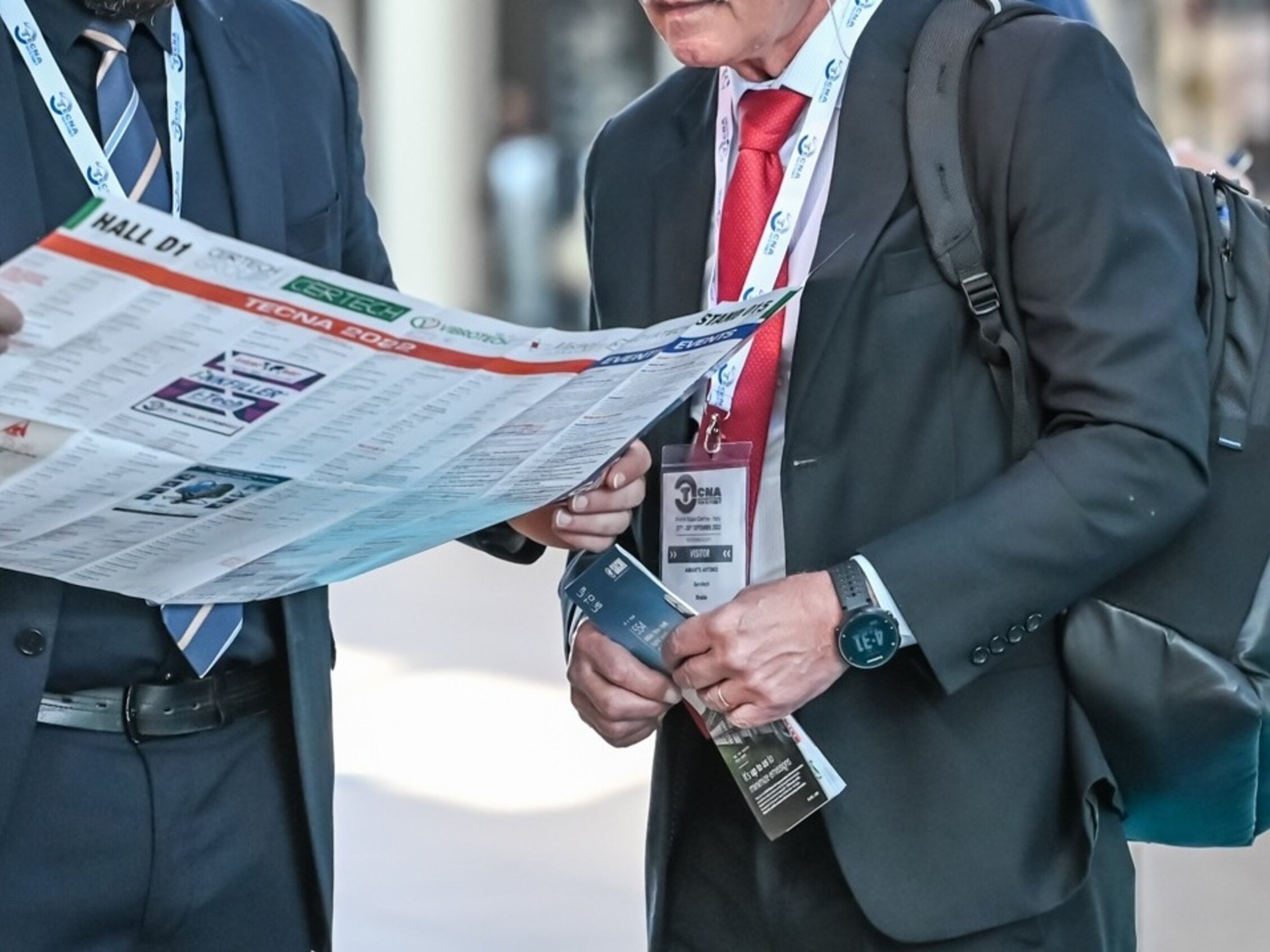 Visitatori che consultano la mappa della fiera durante un evento professionale, con badge al collo e materiali informativi.