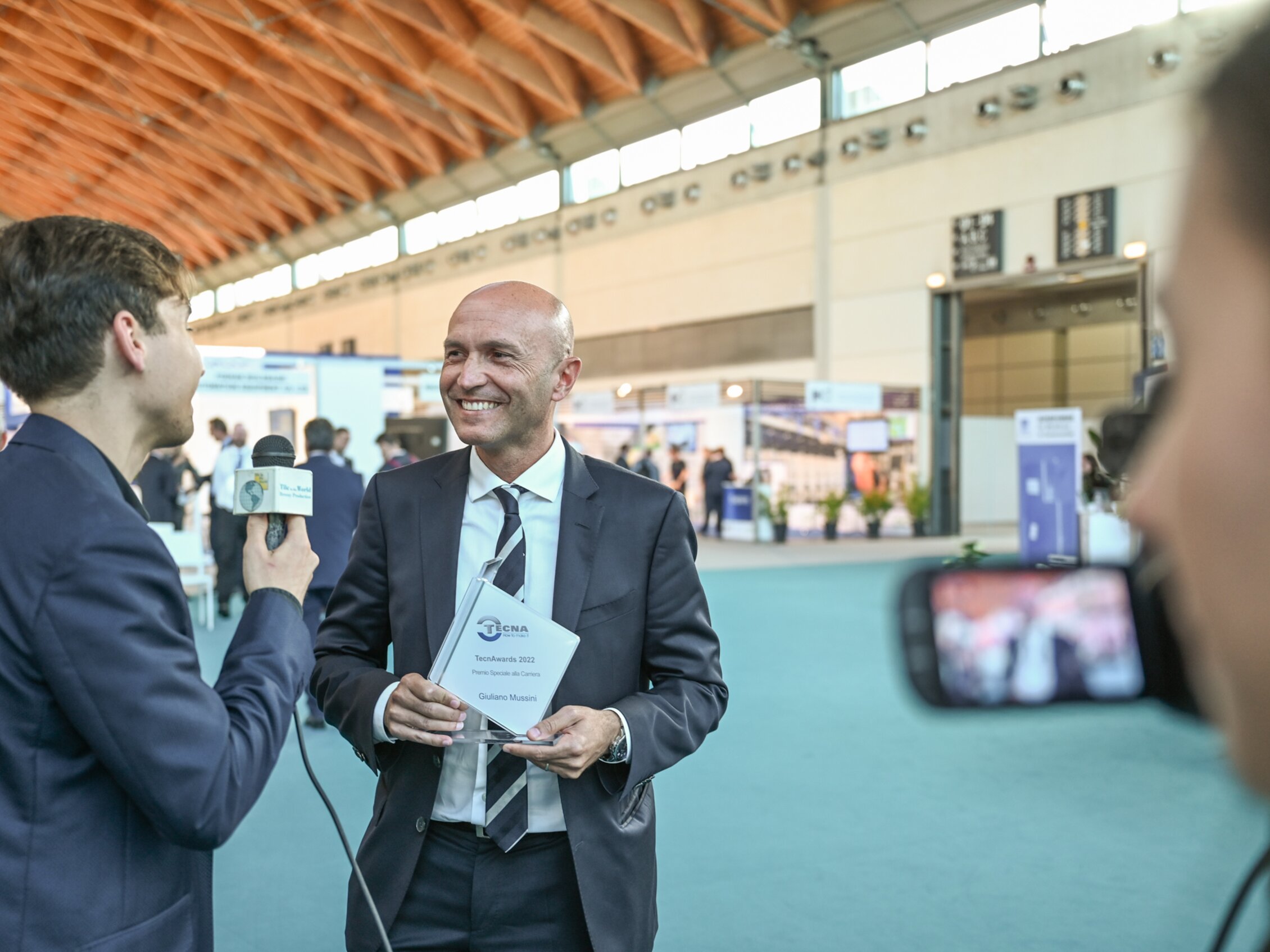 Giornalista intervista un uomo in giacca e cravatta in uno spazio espositivo, con stand e partecipanti sullo sfondo sfocato