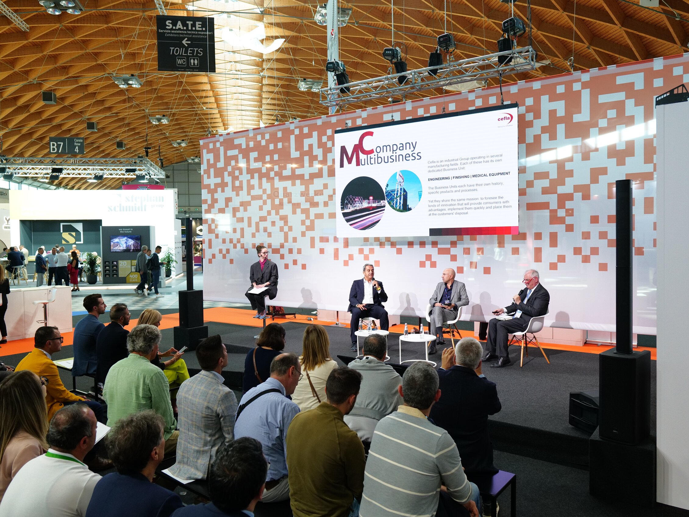 Pubblico seduto ad ascoltare una tavola rotonda in fiera, con quattro relatori sul palco davanti a un grande schermo che mostra la presentazione dell’azienda.