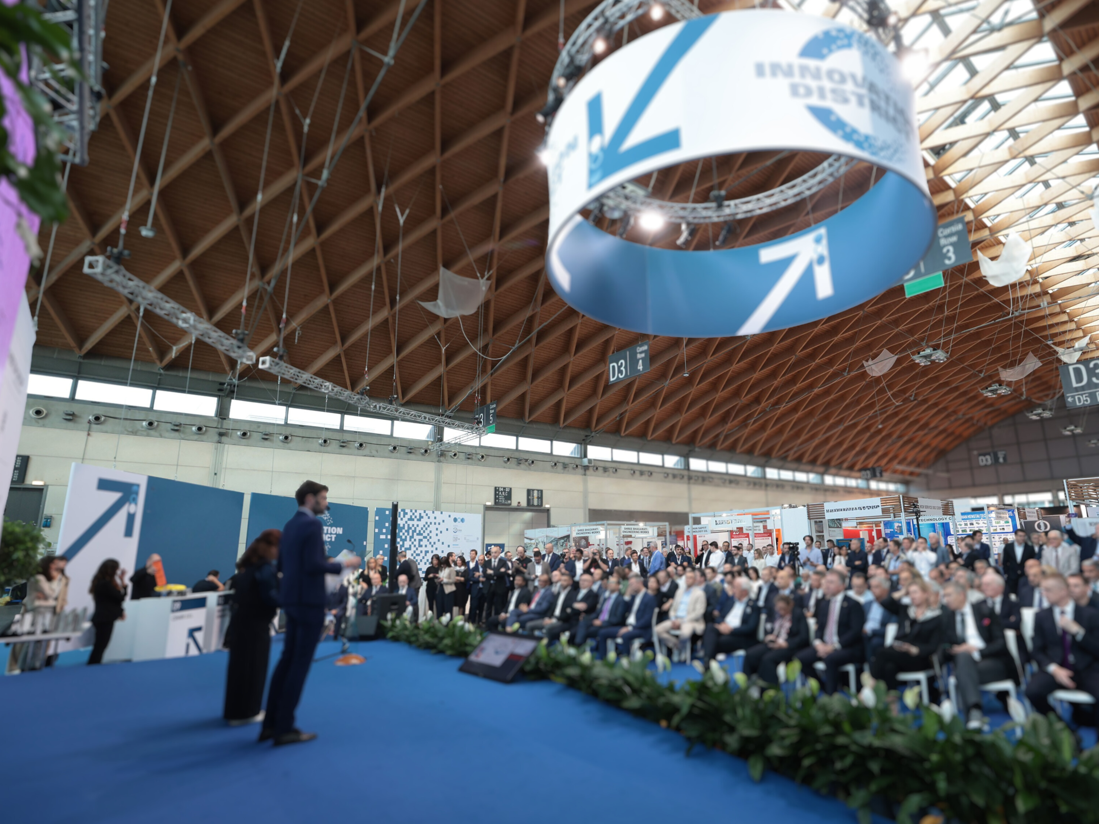 Platea gremita durante una conferenza a Tecna 2024, con il pubblico seduto di fronte al palco blu dove alcuni relatori stanno parlando sotto un grande anello sospeso con grafica dell’evento.​